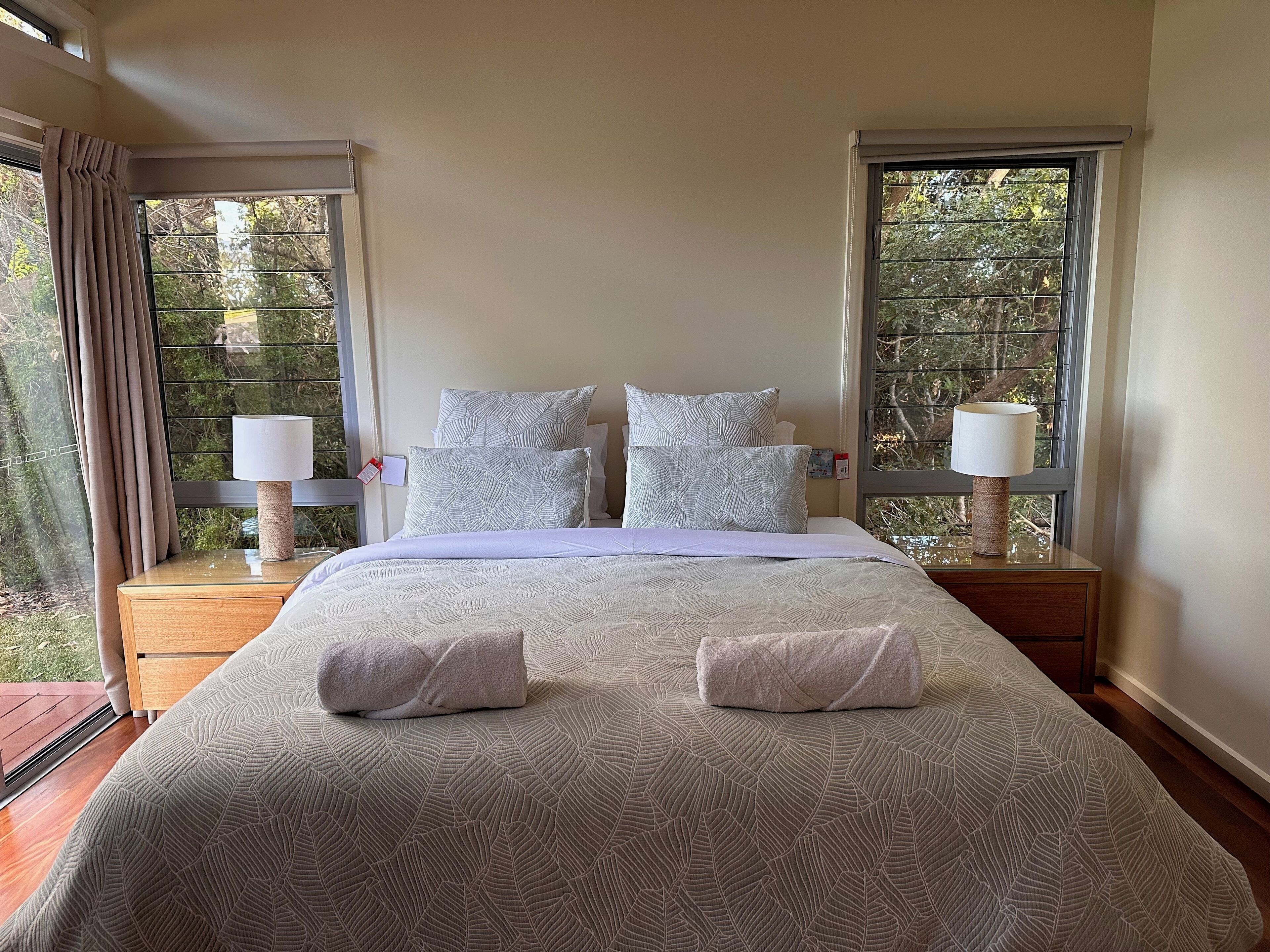A neatly made bed with a leaf-patterned bedspread, two bedside lamps, and a view of trees through the windows.
