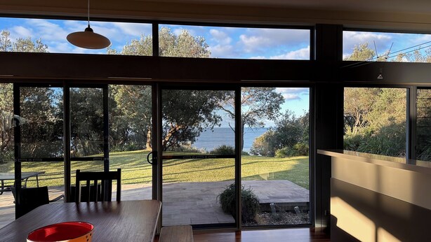 A modern interior with large windows, a wooden dining table, and a red bowl. - Coast@Culburra -  newly renovated  - luxurious beachfront house with spa (Culburra)