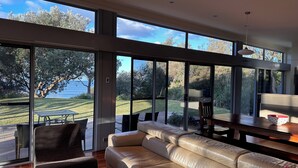 A modern living room with a leather sofa, a glass coffee table, and a dining table. Large windows offer a view of a garden and the sea. - Coast@Culburra -  newly renovated  - luxurious beachfront house with spa (Culburra)