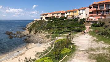 A coastal residential area with a sandy beach, rocky shoreline, and a pathway leading to a row of multi-story buildings with balconies.