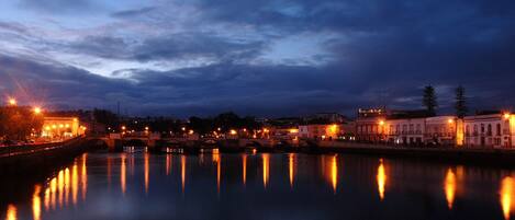 Une vue nocturne d’un paysage urbain au bord de la rivière, avec des bâtiments illuminés dont les reflets se mirent dans l’eau.