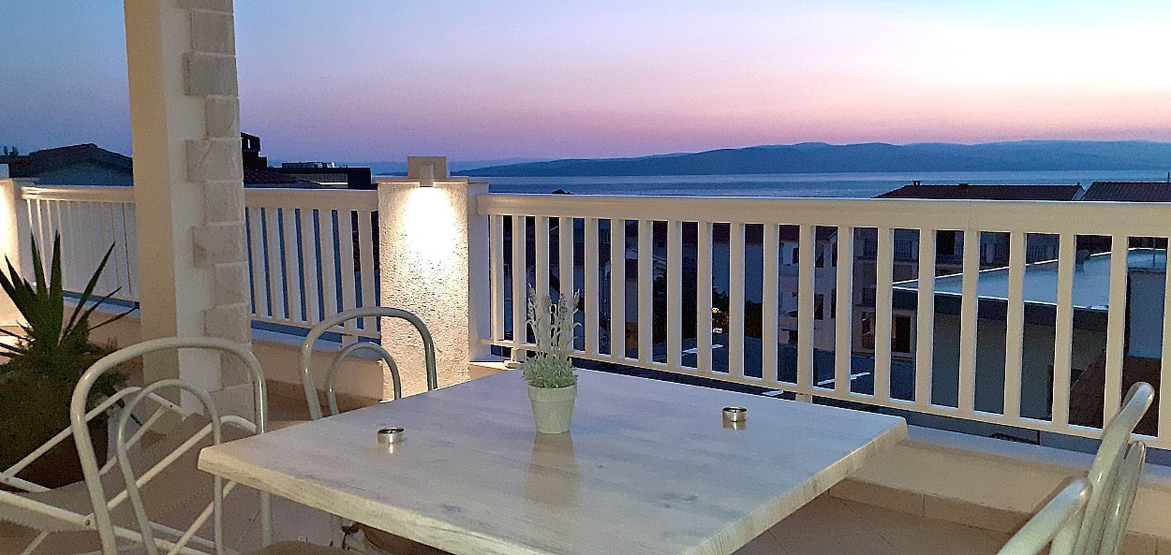A balcony with a table and chairs, overlooking a body of water and distant hills.