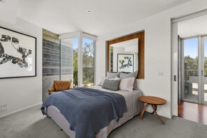 A modern bedroom with a large bed, a side table, and a view of the outdoors through a sliding glass door.