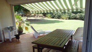 An outdoor patio with a table and chairs, a covered area, and a view of a grassy yard with trees and a fence. - The Desired Creole House (Sainte Anne)