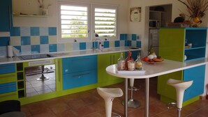 A kitchen with a central island, bar stools, a refrigerator, and a window with blinds. - The Desired Creole House (Sainte Anne)