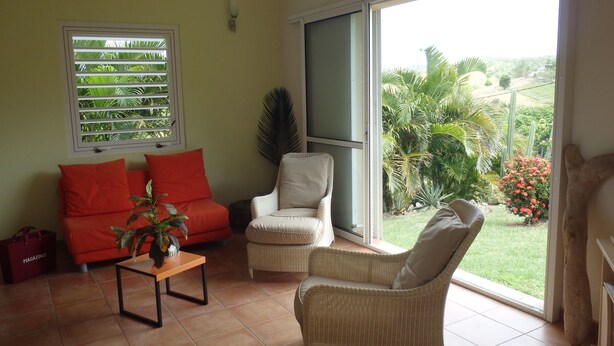 A living room with a sofa, armchair, and a small table. A window with blinds and a view of greenery outside. - The Desired Creole House (Sainte Anne)