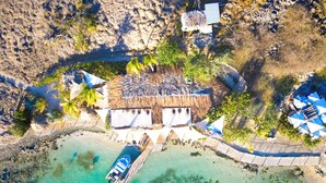 Une vue aérienne d’un complexe hôtelier situé en bord de mer, avec un bâtiment à toit de chaume, des eaux turquoise et limpides, une jetée avec un bateau et plusieurs parasols.