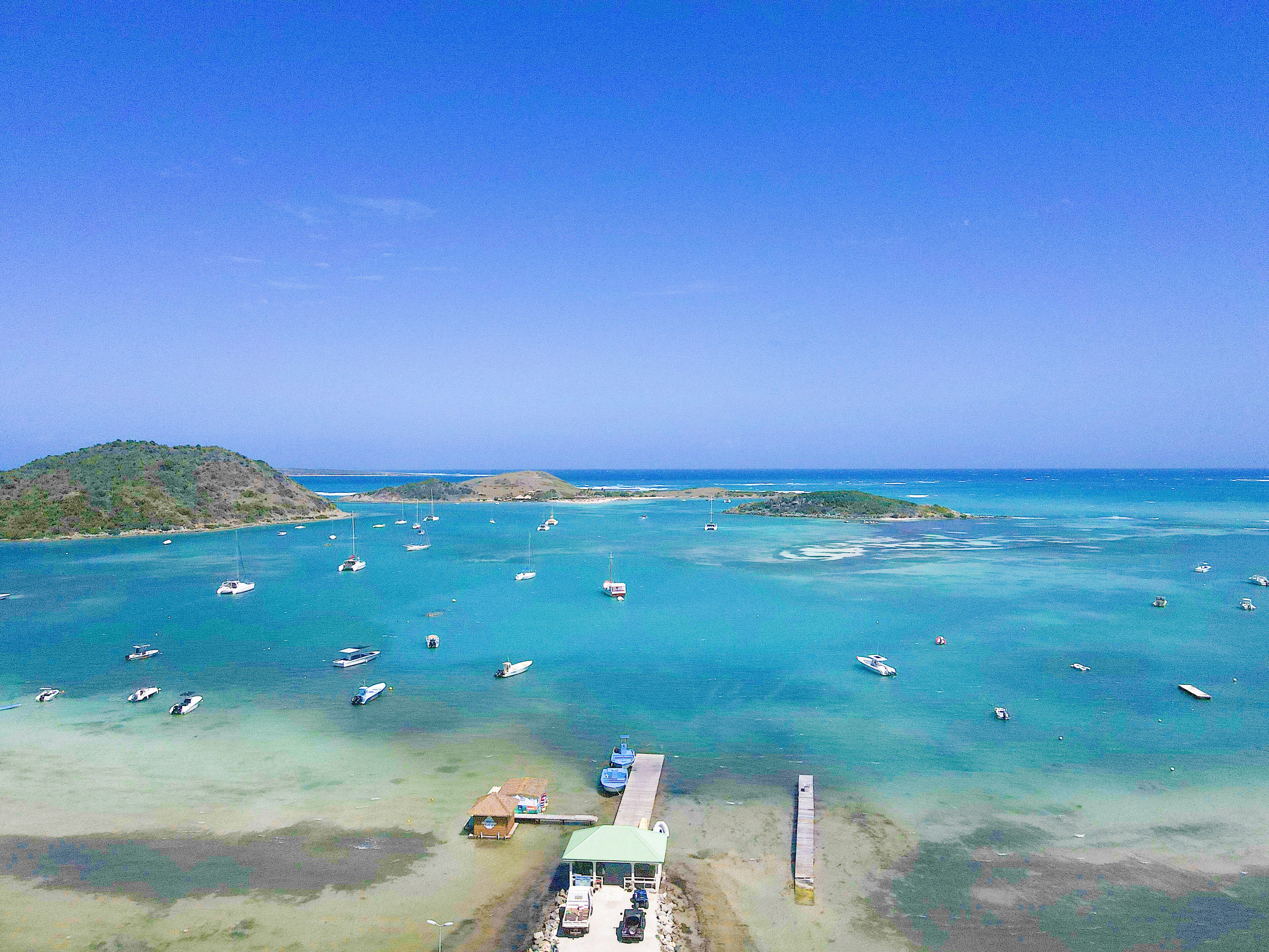 An aerial view of a coastal area with numerous boats anchored in clear blue waters, a small dock with a building, and a hilly landscape in the background.