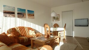 A living room with a sofa, coffee table, and a dining table with chairs. There are two wall-mounted paintings and a television on the wall. - Vacation apartment with a view of the North Sea (Schleswig Holstein)
