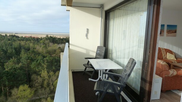 A balcony with a table and chairs, a sofa, and a view of a forested area and beach. - Vacation apartment with a view of the North Sea (Schleswig Holstein)