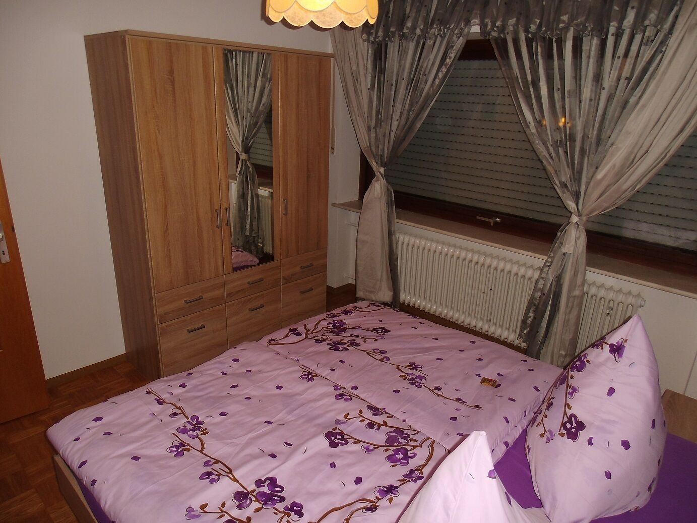 Une chambre avec une armoire en bois, un lit recouvert d’une literie à motifs floraux violets et une fenêtre avec des rideaux.