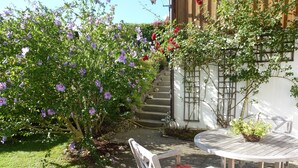 Um jardim com um banco de madeira e uma mesa, cercado por plantas floridas e um edifício branco com escadaria de madeira.