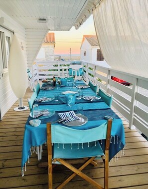 Uma longa mesa de jantar posta com toalhas azuis e cadeiras, em um deck de madeira com guarda-corpo branco e vista para o pôr do sol.