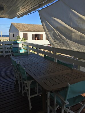 Um deck de madeira ao ar livre com uma mesa e cadeiras, uma casa branca ao fundo e vista para a praia.