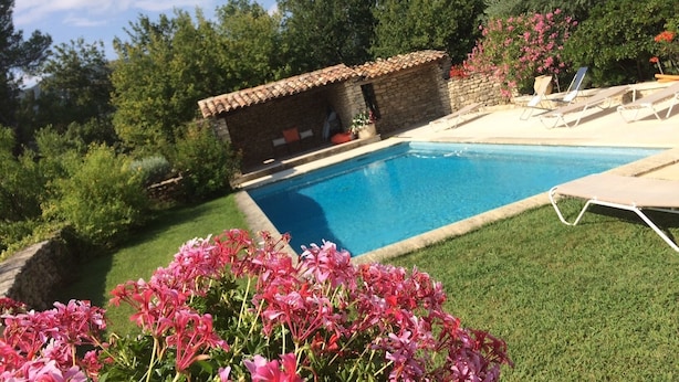 Ein Schwimmbecken mit einem dahinterliegenden Steinhaus, umgeben von Grünpflanzen und Blumen.