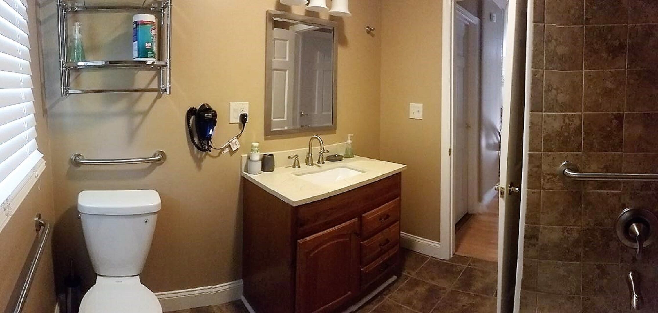 A bathroom with a toilet, sink, and shower. There is a mirror above the sink and a shelf with toiletries.