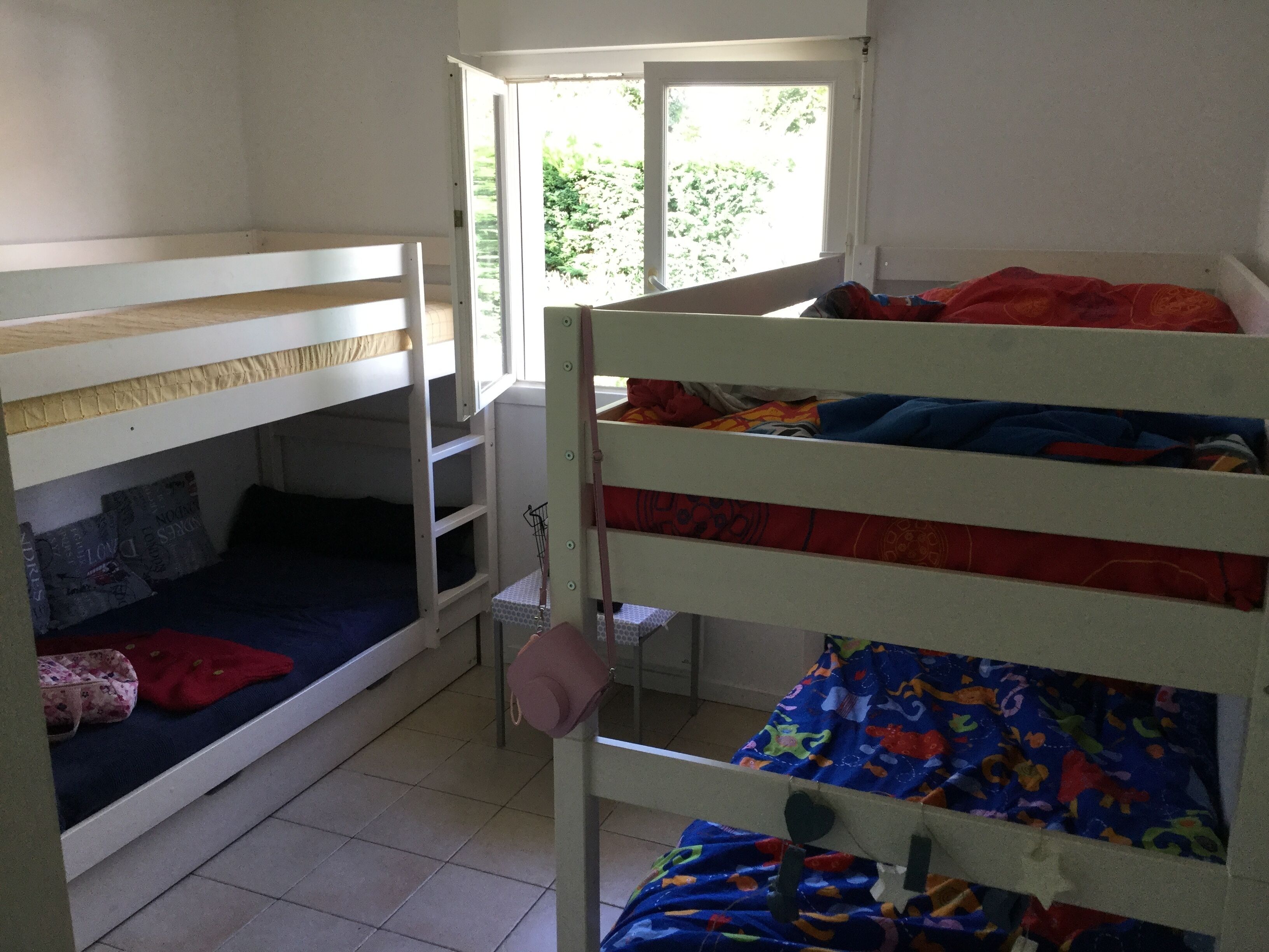 A dormitory room with bunk beds, a small table with a chair, and a window with a view of greenery.