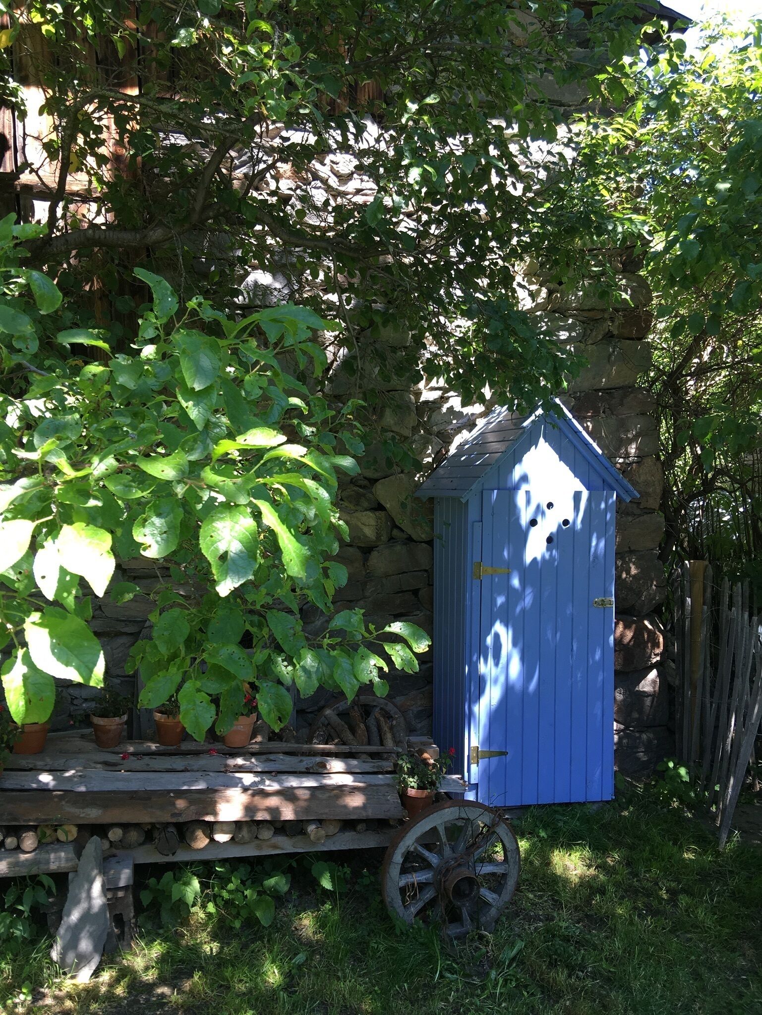 Un jardin avec une charrette en bois remplie de bûches et de plantes en pot, un abri bleu et, au fond, un mur de pierres.