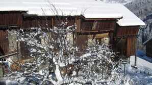 Uma cabana de madeira com telhado coberto de neve, rodeada por árvores e montanhas também cobertas de neve ao fundo.
