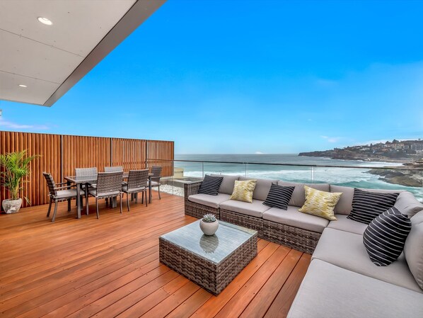 Eine moderne Terrasse im Freien mit Holzdeck, einer Sitzecke mit Kissen, einem Tisch und Blick auf den Ozean.