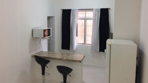 Een kleine, schone kamer met een kitchenette, twee zwarte stoelen, een witte koelkast en een raam met gordijnen.