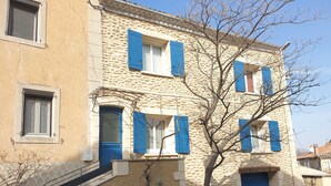 Ein zweistöckiges Gebäude mit Steinmauern und blauen Fensterläden, eine Treppe führt zum Eingang, und ein kleiner Gartenbereich.