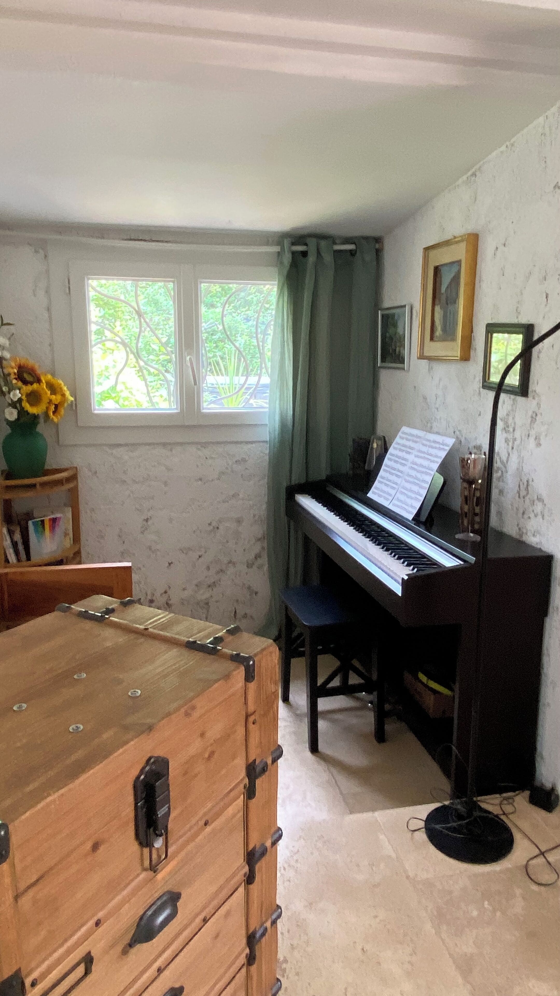Una habitación con un piano, un cofre de madera, una ventana con cortinas y una pared decorada con cuadros.
