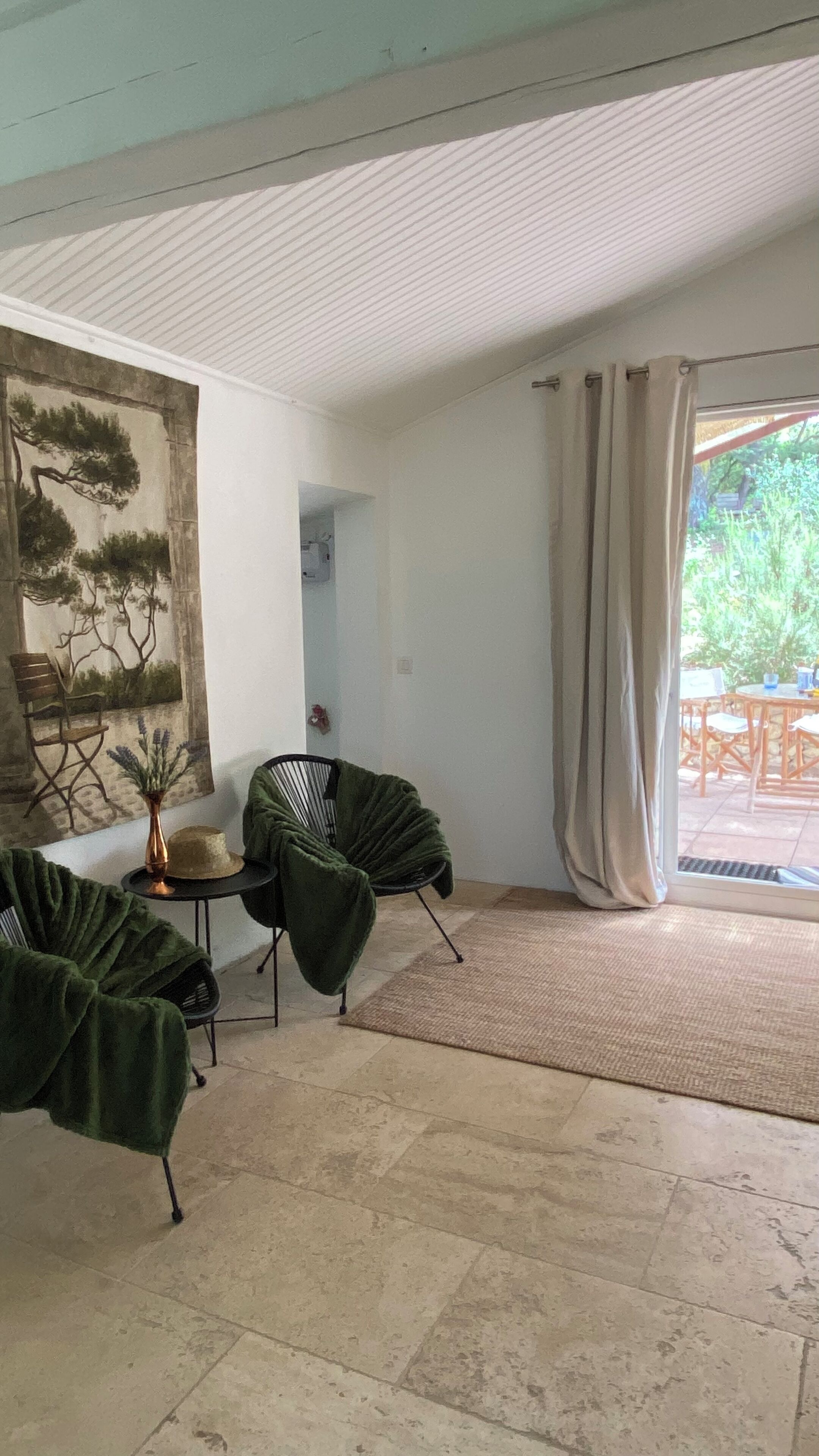 Ein Zimmer mit großem Fenster, einem grünen Sessel, einem kleinen Tisch mit Vase sowie einem Gemälde an der Wand.