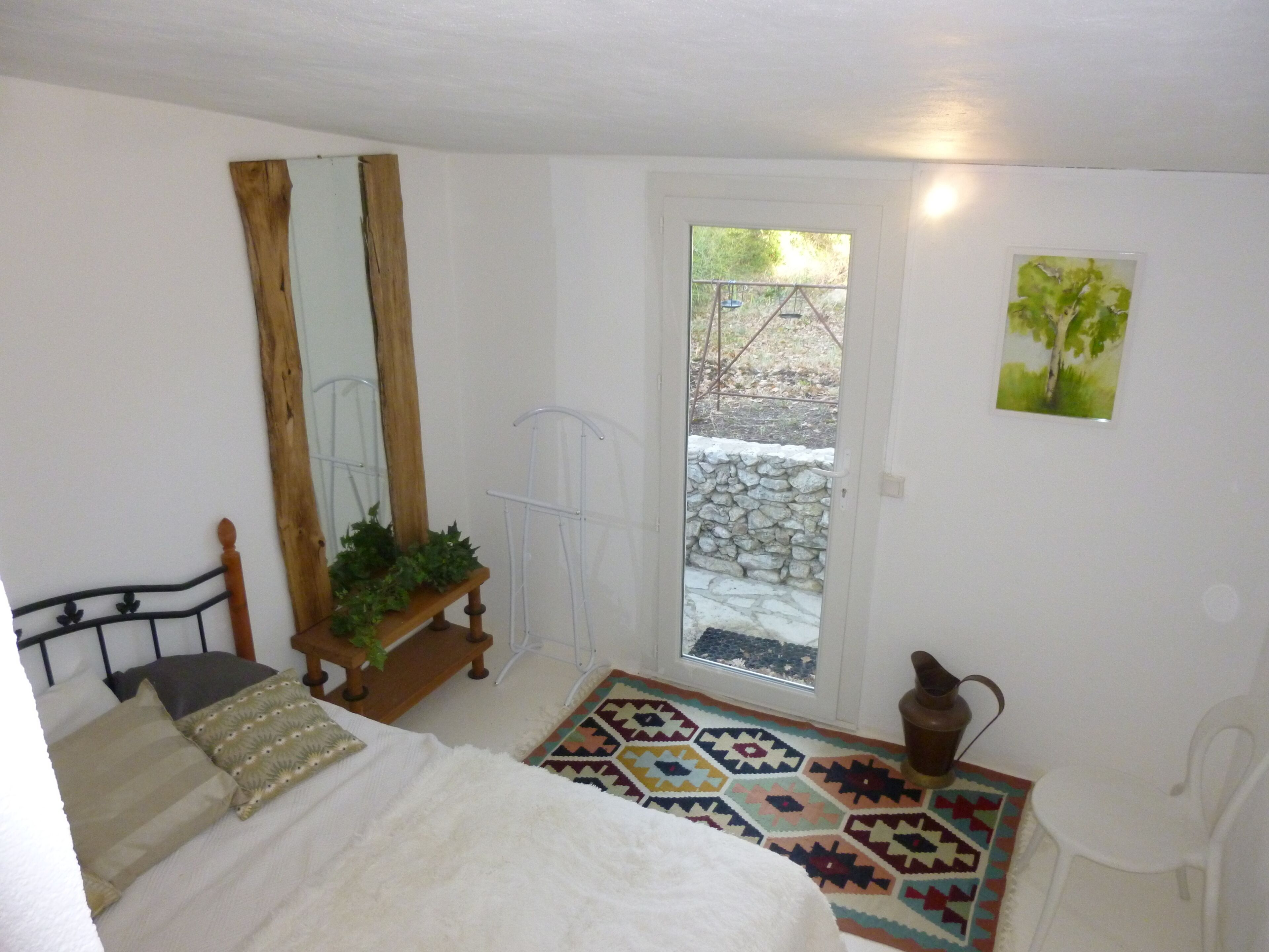 Une chambre avec un lit, un banc en bois agrémenté de plantes, une porte donnant sur l’extérieur, un mur en pierre et un tapis coloré.