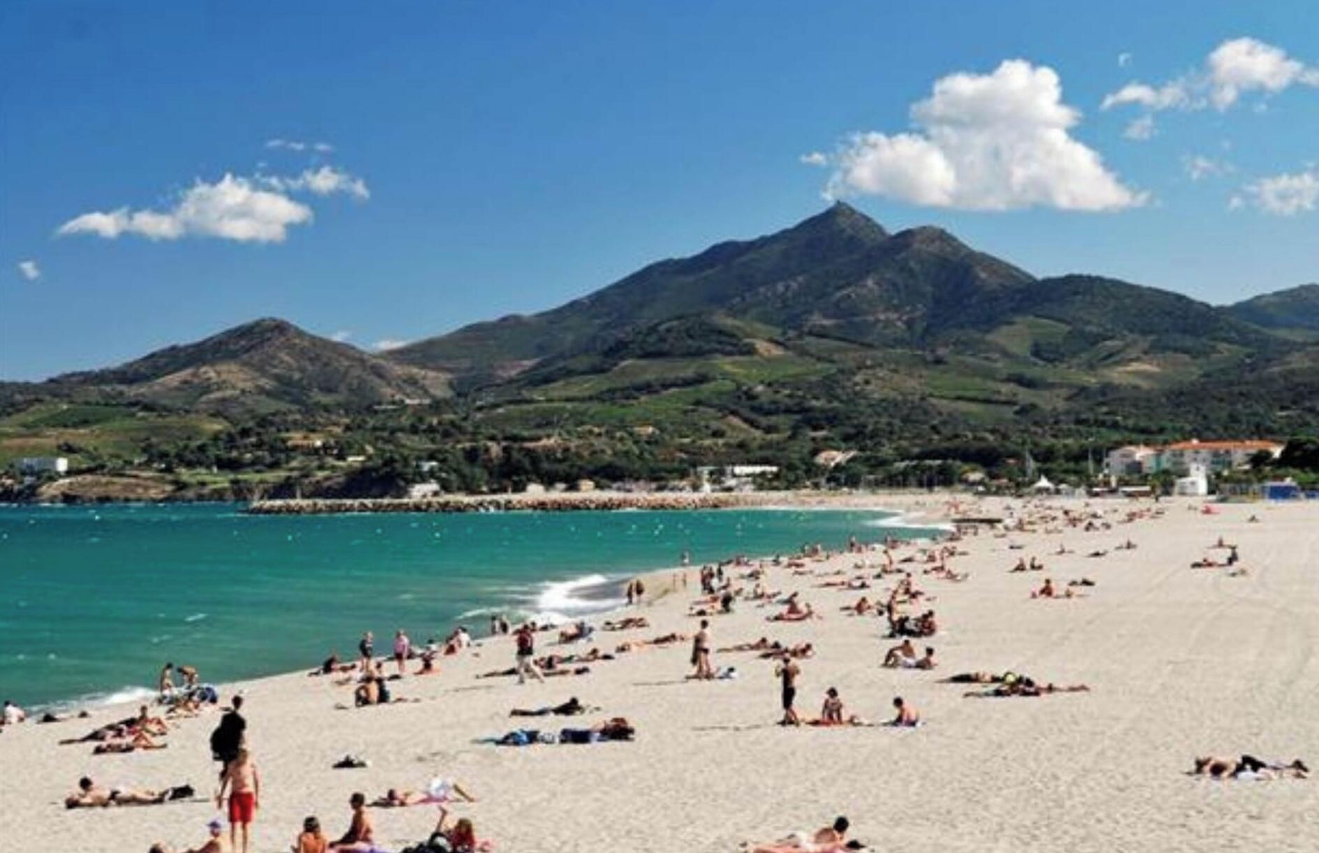 Ein überfüllter Strand mit Menschen, die sich sonnen und schwimmen, umgeben von Bergen und blauem Himmel.