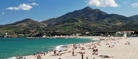 Ein überfüllter Strand mit Menschen, die sich sonnen und schwimmen, umgeben von Bergen und blauem Himmel.