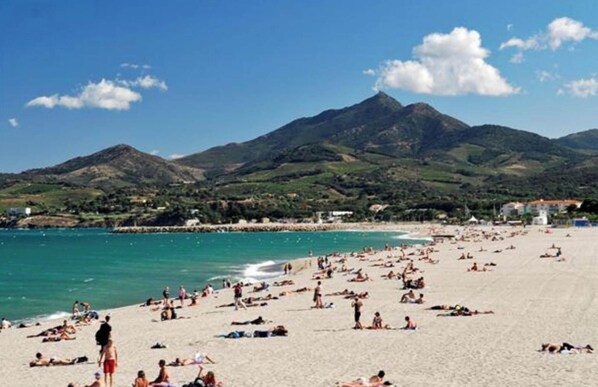 A crowded beach with people sunbathing and swimming, surrounded by mountains and clear blue skies.
