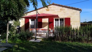 Un edificio de piedra con toldo rojo y contraventanas blancas, rodeado de follaje y un cielo azul despejado.