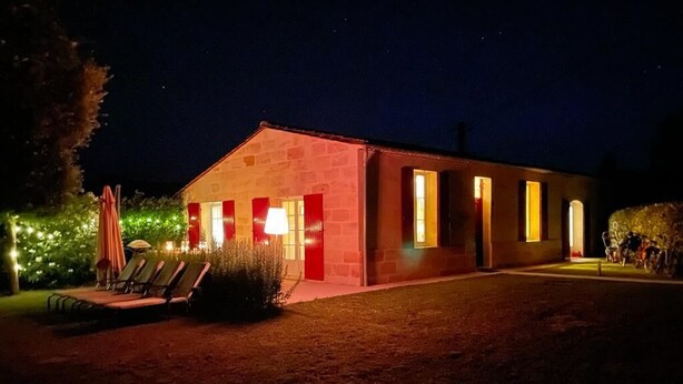Un edificio con puerta y ventanas rojas, rodeado de un jardín con sillas y una sombrilla.