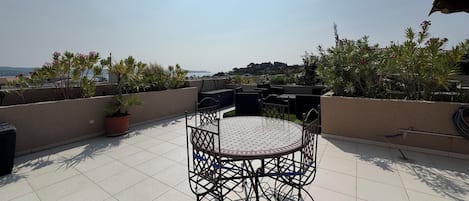 A rooftop terrace with a round table and chairs, surrounded by potted plants and overlooking a scenic view.