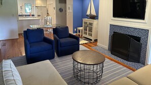 Een woonkamer met een open haard, blauwe fauteuils, een ronde salontafel en een televisie die aan de muur is bevestigd.