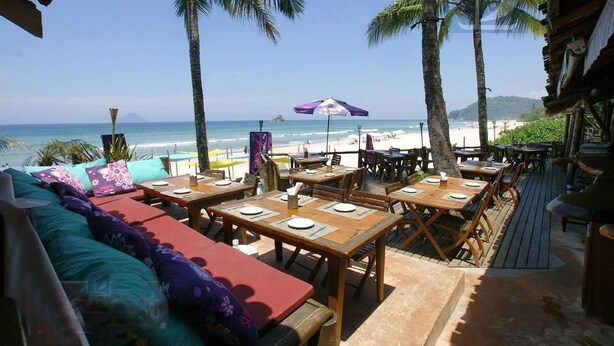 An outdoor dining area with wooden tables and benches, overlooking a beach with palm trees and a clear sky. - Juquehy Condominium Oceanfront 42: best location on the beach  (Praia de Juqueí)