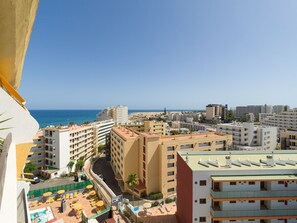 A coastal cityscape with a view of the sea, featuring multi-story buildings, a parking area, and outdoor seating.
