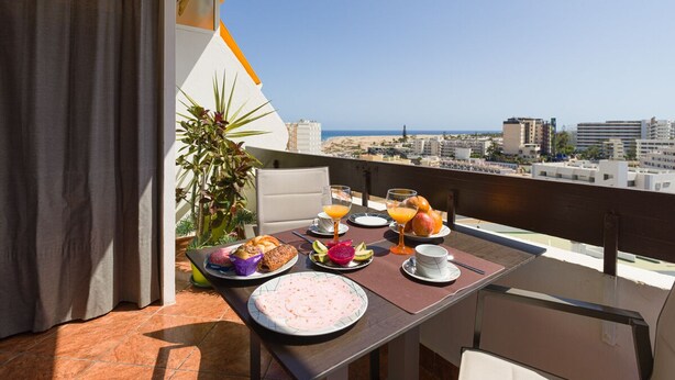 Ein Balkon mit Frühstückstisch, der Blick auf eine Stadtlandschaft und das Meer freigibt.