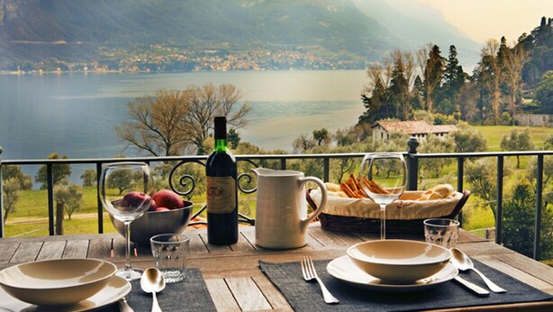 Une table dressée avec de la vaisselle, une bouteille de vin et un panier de pain, donnant sur un lac avec des montagnes en arrière-plan.