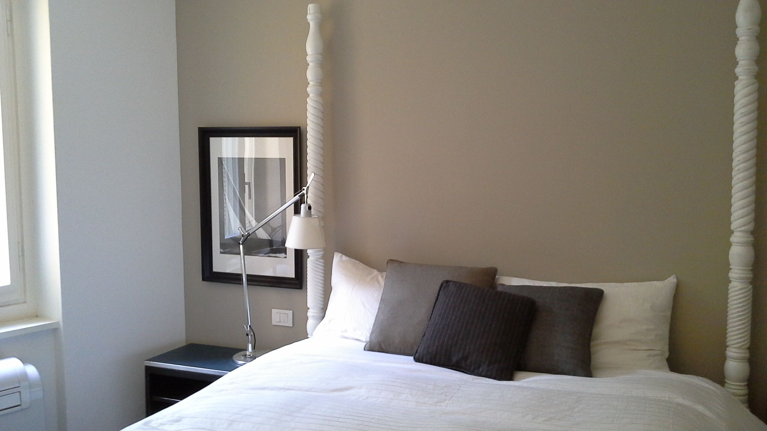 A neatly made bed with white and gray linens, a bedside table with a lamp, and a framed picture on the wall.