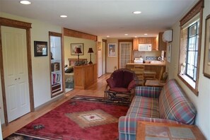 A cozy living room with a red carpet, a plaid sofa, a wicker chair, and a kitchen area with wooden cabinets and a window. - Dragon's Lair / Garden Loft, Very Private, Cov. Gar. 1 BR. 1 BA. 1 Mi to MS. (Mt Shasta)