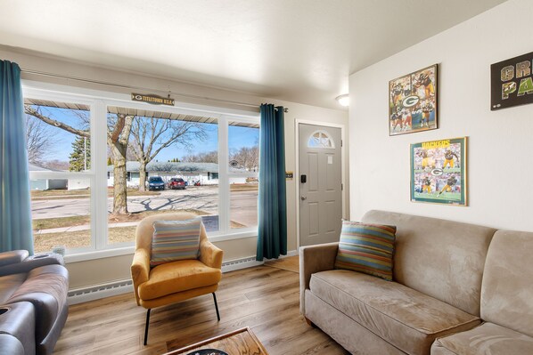 A living room with a sofa, armchair, and a view of a parking lot through large windows. - Classic Oneida Tailgate House - 5 minute walk to Lambeau (Green Bay)