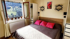 A bedroom with a large bed, a window with a view of a landscape, a wardrobe, and a wall-mounted light fixture. - 68 m² *** self-contained apartment, large terrace, parking, surrounded by mountains. (Haute-Garonne)