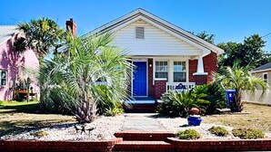 A single-story house with a red brick facade, a blue front door, and a small front yard with palm trees and a potted plant.