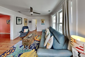 Un salon avec un canapé bleu, un ventilateur de plafond, un tapis à motifs et une fenêtre avec des rideaux.