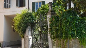 Un edificio con un ingresso sorvegliato da un cancello, un muro in pietra con vegetazione e un cielo sereno.