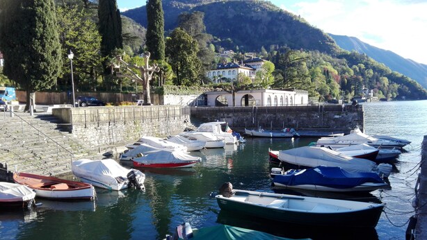 Una marina con diverse imbarcazioni ormeggiate, un muro in pietra e un edificio con finestre ad arco sullo sfondo.