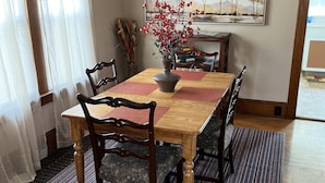 Un comedor con una mesa de madera, sillas, un jarrón con flores rojas, un cuadro en la pared y una ventana con cortinas traslúcidas.