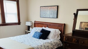 Un dormitorio con una cama de madera, una mesita de noche con lámpara, un aparador y un cuadro en la pared.